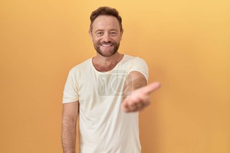 Foto de Hombre de mediana edad con barba de pie sobre fondo amarillo sonriente alegre ofreciendo la palma de la mano dando asistencia y aceptación. - Imagen libre de derechos