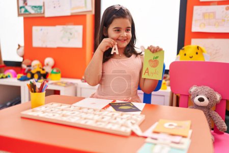 Téléchargez les photos : Adorable fille hispanique jouant avec le jeu de puzzle de vocabulaire assis sur la table à la maternelle - en image libre de droit