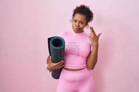 Photo for Young hispanic woman with curly hair holding yoga mat over pink background shooting and killing oneself pointing hand and fingers to head like gun, suicide gesture. - Royalty Free Image