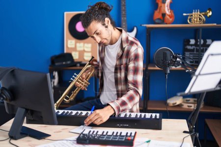 Foto de Young hispanic man musician composing song holding trumpet at music studio - Imagen libre de derechos