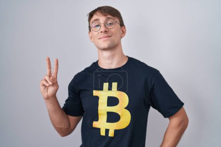 Foto de Hombre rubio caucásico con camiseta bitcoin sonriendo mirando a la cámara mostrando los dedos haciendo señal de victoria. número dos. - Imagen libre de derechos