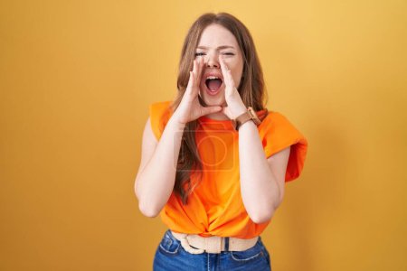 Photo for Caucasian woman standing over yellow background shouting angry out loud with hands over mouth - Royalty Free Image