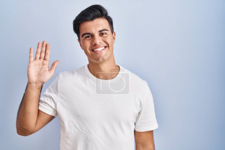 Téléchargez les photos : Homme hispanique debout sur fond bleu renonçant à dire bonjour heureux et souriant, geste de bienvenue amical - en image libre de droit