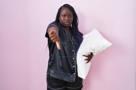 Téléchargez les photos : Jeune femme africaine portant un pyjama étreignant oreiller regardant malheureux et en colère montrant rejet et négatif avec les pouces vers le bas geste. mauvaise expression. - en image libre de droit