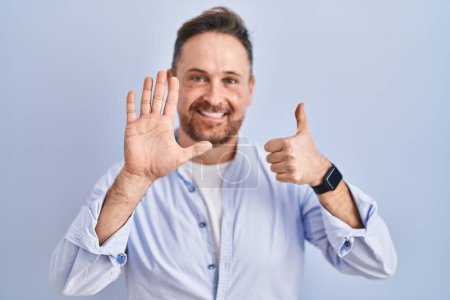 Foto de Hombre caucásico de mediana edad de pie sobre fondo azul mostrando y apuntando hacia arriba con los dedos número seis mientras sonríe confiado y feliz. - Imagen libre de derechos