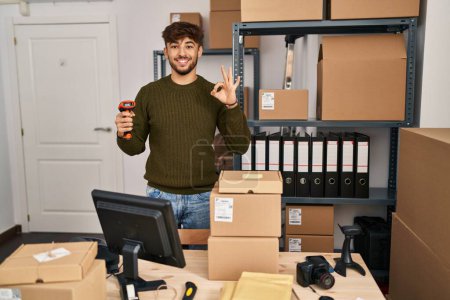 Foto de Hombre árabe con barba trabajando en el comercio de la pequeña empresa sosteniendo el escáner de código de barras haciendo signo bien con los dedos, sonriendo gesto amistoso excelente símbolo - Imagen libre de derechos
