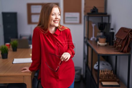 Foto de Young redhead woman business worker smiling confident holding glasses at office - Imagen libre de derechos