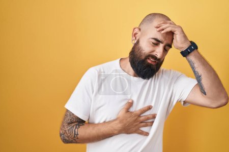 Téléchargez les photos : Jeune homme hispanique avec barbe et tatouages debout sur fond jaune touchant le front pour la maladie et la fièvre, grippe et rhume, virus malade - en image libre de droit
