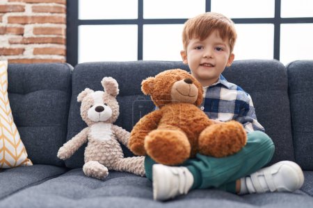 Photo for Adorable toddler holding teddy bear sitting on sofa at home - Royalty Free Image