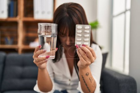 Foto de Joven mujer hispana hermosa paciente estresada sosteniendo pastillas tratamiento y vaso de agua en la clínica de psicología - Imagen libre de derechos