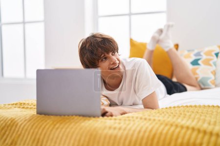 Téléchargez les photos : Jeune homme blond utilisant un ordinateur portable couché sur le lit dans la chambre - en image libre de droit