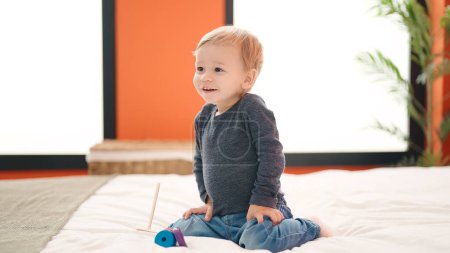 Téléchargez les photos : Adorable tout-petit blond jouant avec des cerceaux jouet assis sur le lit dans la chambre - en image libre de droit