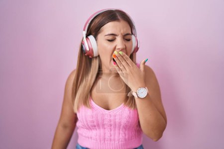 Foto de Mujer rubia joven escuchando música usando auriculares aburridos bostezando cansada cubriendo la boca con la mano. inquietud y somnolencia. - Imagen libre de derechos