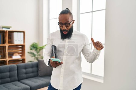 Foto de Hombre afroamericano que trabaja en la oficina de terapia con teléfono inteligente asustado y sorprendido con la boca abierta para sorpresa, cara de incredulidad - Imagen libre de derechos