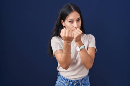 Téléchargez les photos : Young hispanic woman standing over blue background ready to fight with fist defense gesture, angry and upset face, afraid of problem - en image libre de droit