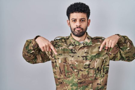 Téléchargez les photos : Homme arabe portant uniforme de camouflage de l'armée pointant vers le bas regardant triste et contrarié, indiquant la direction avec les doigts, malheureux et déprimé. - en image libre de droit