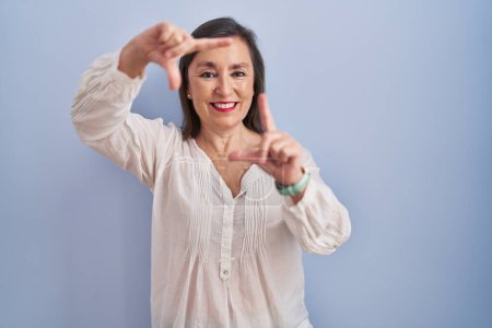 Foto de Mujer hispana de mediana edad de pie sobre fondo azul sonriendo haciendo marco con las manos y los dedos con la cara feliz. concepto de creatividad y fotografía. - Imagen libre de derechos