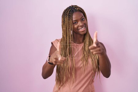 Foto de African american woman with braided hair standing over pink background pointing fingers to camera with happy and funny face. good energy and vibes. - Imagen libre de derechos