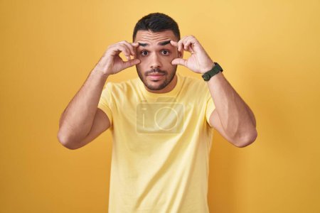 Téléchargez les photos : Jeune homme hispanique debout sur fond jaune essayant d'ouvrir les yeux avec les doigts, somnolent et fatigué pour la fatigue matinale - en image libre de droit