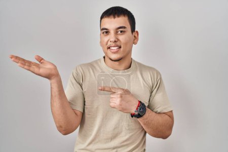 Téléchargez les photos : Jeune homme arabe portant t-shirt décontracté étonné et souriant à la caméra tout en présentant avec la main et pointant du doigt. - en image libre de droit