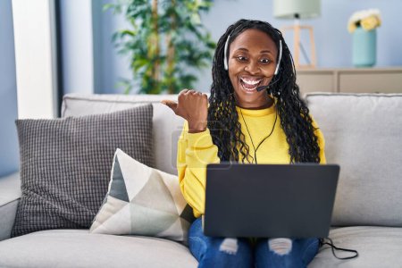 Photo for African woman working at home wearing operator headset pointing thumb up to the side smiling happy with open mouth - Royalty Free Image