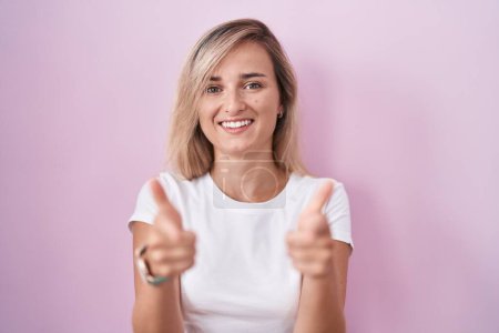 Téléchargez les photos : Young blonde woman standing over pink background pointing fingers to camera with happy and funny face. good energy and vibes. - en image libre de droit