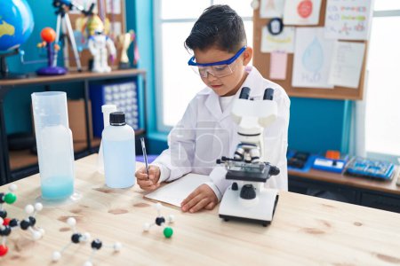 Téléchargez les photos : Adorable élève hispanique garçon en utilisant des notes de microscope écriture à la salle de classe de laboratoire - en image libre de droit