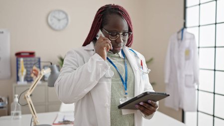 Téléchargez les photos : Femme africaine avec médecin cheveux tressés parlant sur smartphone lecture de la tablette à la clinique - en image libre de droit
