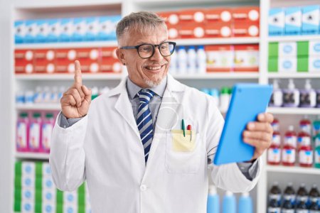 Foto de Hombre hispano con cabello gris trabajando en farmacia haciendo videollamada con tableta sonriendo con una idea o pregunta señalando con el dedo con la cara feliz, número uno - Imagen libre de derechos