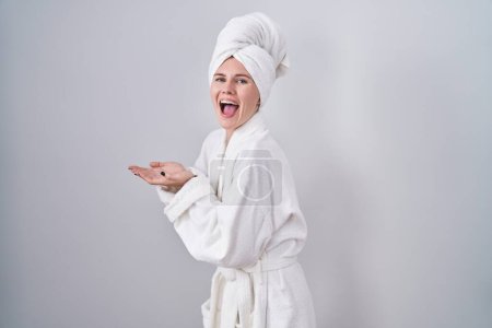 Foto de Mujer rubia caucásica usando albornoz señalando a un lado con las manos abiertas las palmas mostrando espacio de copia, presentando anuncio sonriendo emocionada feliz - Imagen libre de derechos