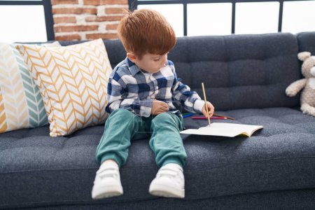 Foto de Adorable dibujo infantil en el cuaderno sentado en el sofá en casa - Imagen libre de derechos