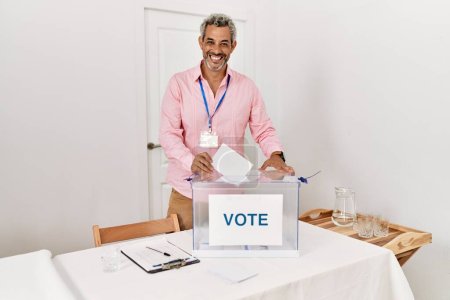 Téléchargez les photos : Moyen-âge homme aux cheveux gris président de table électorale mettant le vote sur les urnes au collège électoral - en image libre de droit