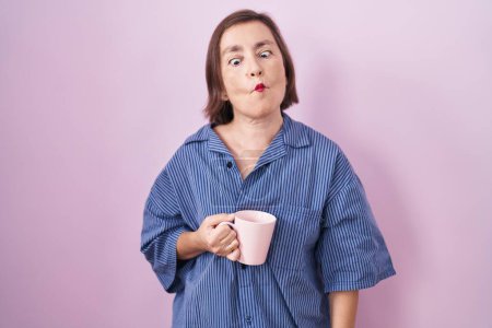 Téléchargez les photos : Femme hispanique du Moyen Age boire une tasse de café faire visage de poisson avec des lèvres, geste fou et comique. expression drôle. - en image libre de droit