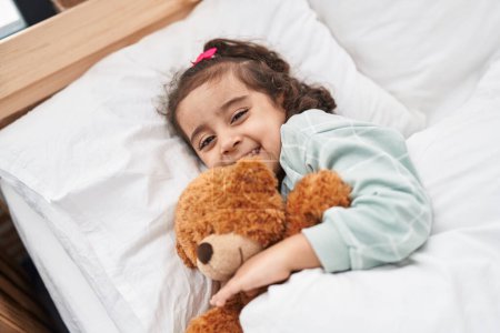 Photo for Adorable hispanic girl hugging teddy bear lying on bed at bedroom - Royalty Free Image