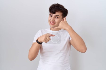 Téléchargez les photos : Jeune homme non binaire portant un t-shirt blanc décontracté souriant faisant parler au téléphone geste et vous pointant du doigt. Appelle-moi. - en image libre de droit