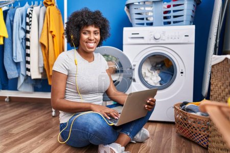 Foto de Mujer afroamericana escuchando música esperando la lavadora en la lavandería - Imagen libre de derechos
