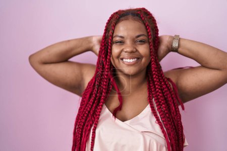 Téléchargez les photos : Femme afro-américaine avec des cheveux tressés debout sur fond rose relaxant et étirant, bras et mains derrière la tête et le cou souriant heureux - en image libre de droit