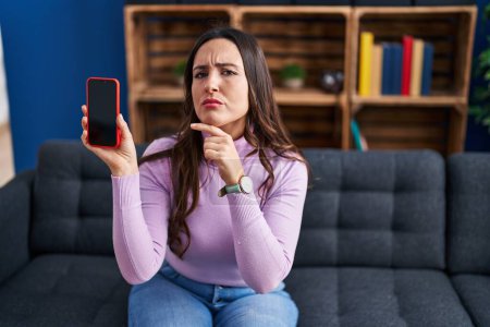 Photo for Young brunette woman holding smartphone showing blank screen serious face thinking about question with hand on chin, thoughtful about confusing idea - Royalty Free Image