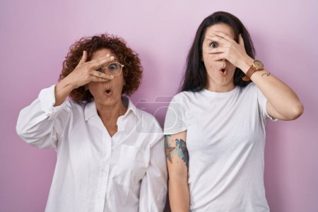 Foto de Madre e hija hispanas vistiendo una camiseta blanca casual sobre fondo rosa asomándose en conmoción cubriendo cara y ojos con la mano, mirando a través de los dedos con expresión avergonzada. - Imagen libre de derechos