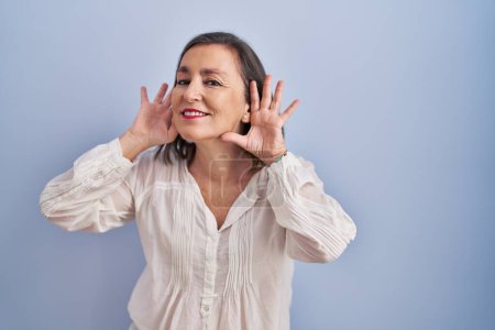 Photo for Middle age hispanic woman standing over blue background trying to hear both hands on ear gesture, curious for gossip. hearing problem, deaf - Royalty Free Image