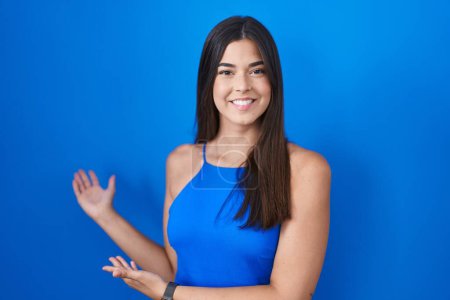 Foto de Hispanic woman standing over blue background inviting to enter smiling natural with open hand - Imagen libre de derechos