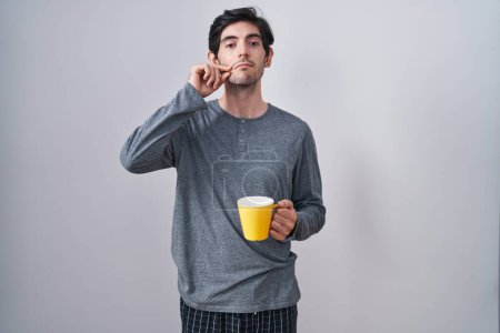 Téléchargez les photos : Jeune homme hispanique portant un pyjama boire une tasse de bouche de café et les lèvres fermées comme zip avec les doigts. secret et silencieux, tabou parlant - en image libre de droit