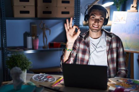 Photo for Young hispanic man sitting at art studio with laptop late at night smiling positive doing ok sign with hand and fingers. successful expression. - Royalty Free Image