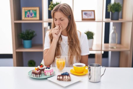 Foto de Mujer caucásica joven comiendo pasteles t para el desayuno sensación de malestar y tos como síntoma de resfriado o bronquitis. concepto de atención sanitaria. - Imagen libre de derechos