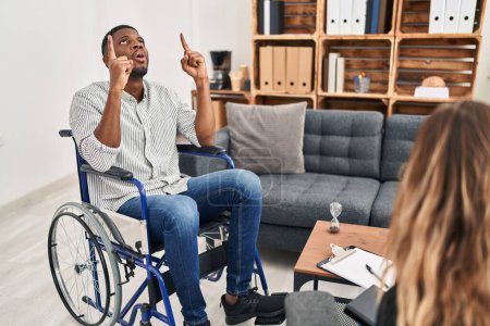 Photo for African american man doing therapy sitting on wheelchair amazed and surprised looking up and pointing with fingers and raised arms. - Royalty Free Image
