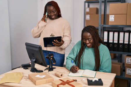 Téléchargez les photos : Femmes afro-américaines commerce électronique travailleurs d'affaires parlant sur smartphone écriture sur ordinateur portable au bureau - en image libre de droit
