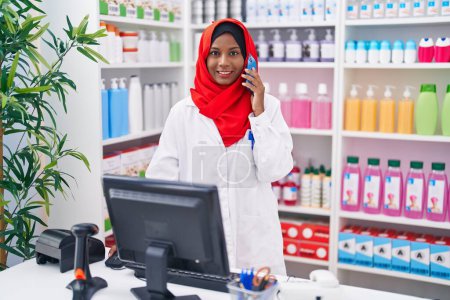 Photo for Young beautiful woman pharmacist talking on smartphone using computer at pharmacy - Royalty Free Image