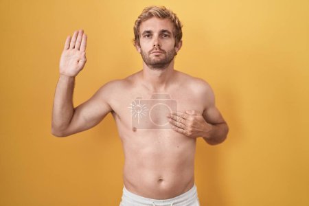Téléchargez les photos : Homme caucasien debout torse nu portant un écran solaire jurant avec la main sur la poitrine et la paume ouverte, faisant un serment de fidélité - en image libre de droit