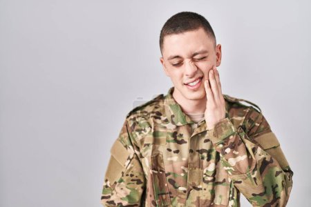 Téléchargez les photos : Jeune homme portant un uniforme de camouflage militaire touchant la bouche avec la main avec une expression douloureuse à cause de maux de dents ou de maladies dentaires sur les dents. dentiste - en image libre de droit