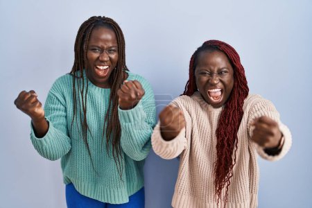 Foto de Dos mujeres africanas de pie sobre fondo azul enojadas y locas levantando puños frustradas y furiosas mientras gritaban con ira. rabia y concepto agresivo. - Imagen libre de derechos